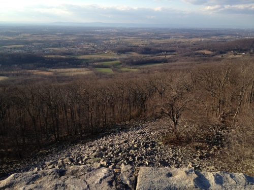 A panoramic view from a rocky overlook, showcasing a valley with sparse trees and distant hills under a cloudy sky.