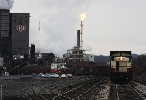 A train on tracks near an industrial site with smokestacks and a flame, set against a cloudy sky.