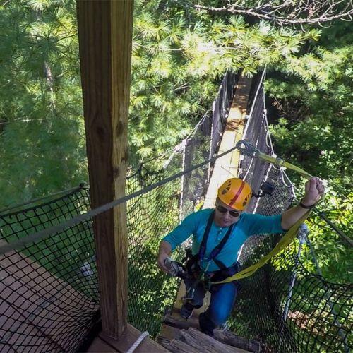 A person in a helmet and harness navigates a rope bridge surrounded by trees in an outdoor adventure setting.