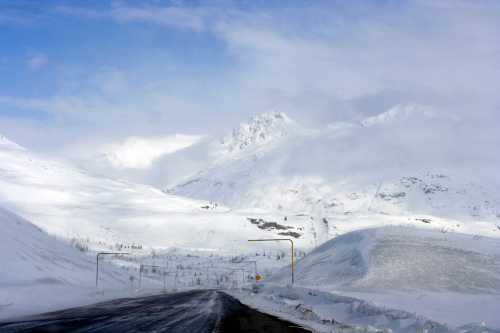 A snowy mountain landscape with a winding road disappearing into the distance under a cloudy sky.