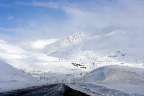 A snowy mountain landscape with a winding road disappearing into the distance under a cloudy sky.