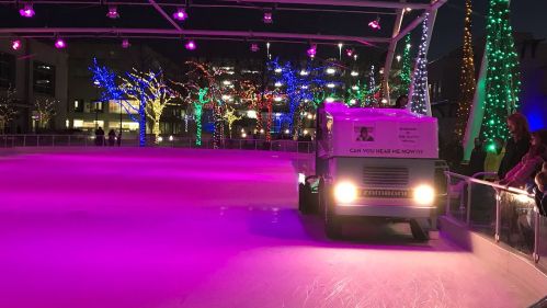 A zamboni cleans an ice rink surrounded by colorful holiday lights and trees at night.