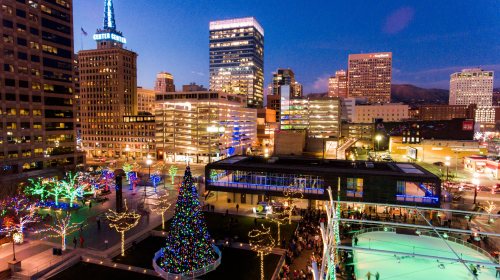 A vibrant city skyline at dusk, featuring colorful holiday lights and a large decorated Christmas tree in a public square.