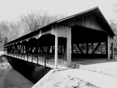 A black and white image of a wooden covered bridge spanning a calm stream, surrounded by bare trees.