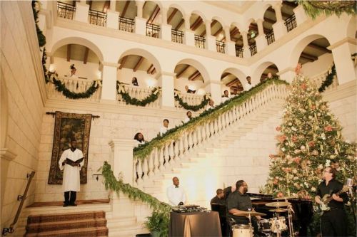 A festive indoor scene with a decorated Christmas tree, musicians, and people gathered on a grand staircase.