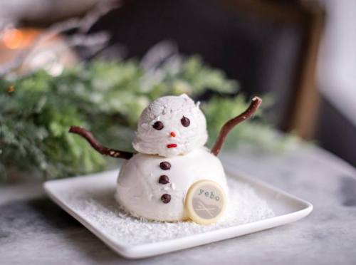 A cute snowman dessert made of ice cream, with pretzel arms, chocolate buttons, and a cookie on a snowy plate.
