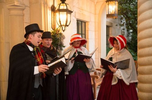 A group of carolers in festive attire singing together under lantern light on a porch.