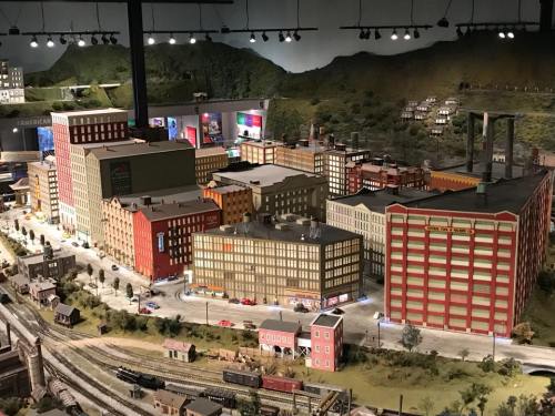 A detailed miniature cityscape featuring colorful buildings, roads, and train tracks, set against a hilly backdrop.