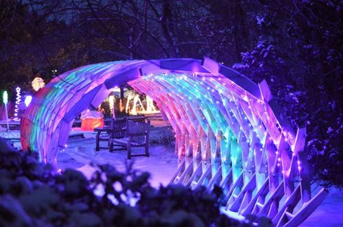 A colorful, illuminated archway in a snowy park, with benches and festive lights in the background.