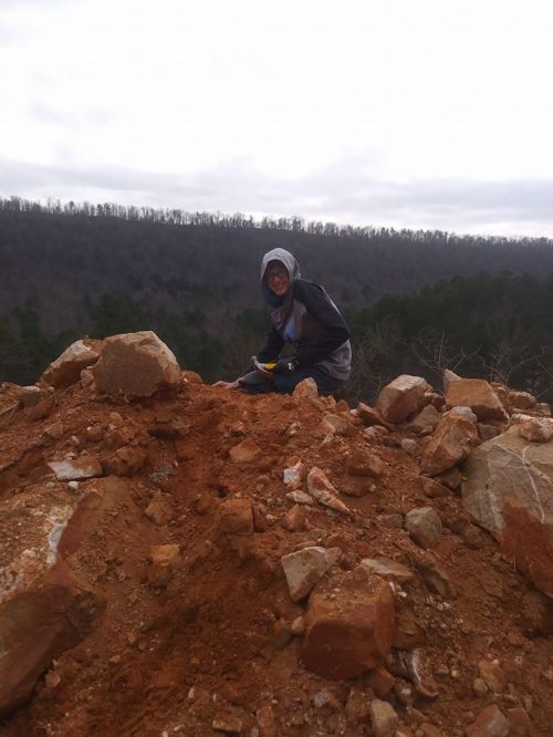 A person in a hoodie sits on a pile of rocks and dirt, with a forested hillside in the background under a cloudy sky.
