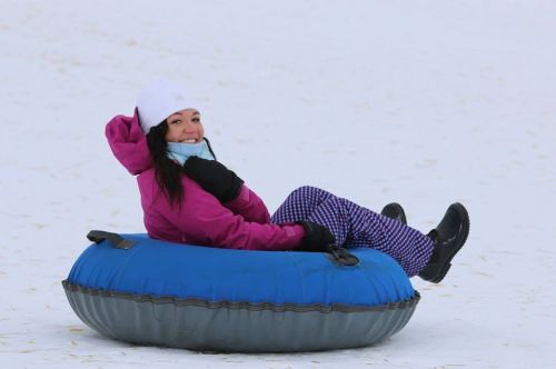 A woman in a pink jacket and white hat sits on a blue snow tube, smiling against a snowy background.