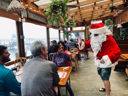 A man in a Santa costume and sunglasses greets diners in a festive restaurant decorated with plants.