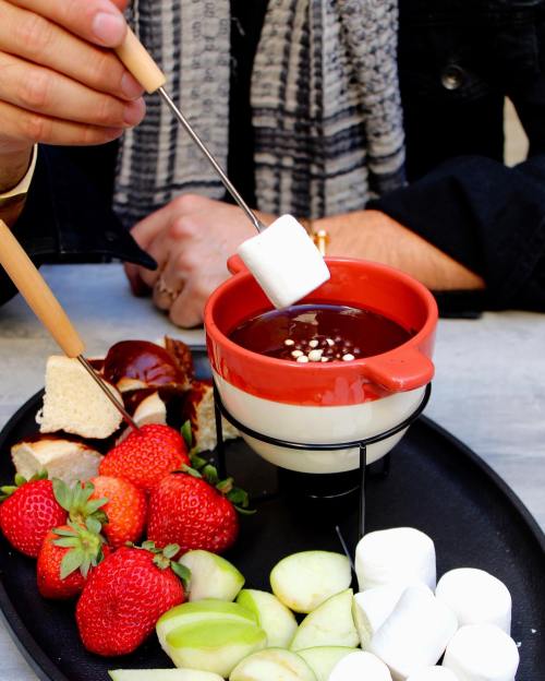 A hand holds a skewer with a marshmallow over a bowl of chocolate fondue, surrounded by fruits and treats on a platter.