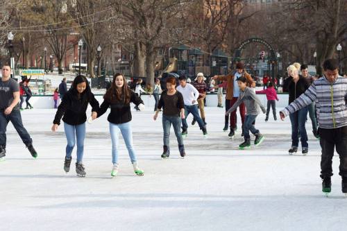 A group of people ice skating on a rink, enjoying a sunny day in a park. Trees and buildings are in the background.
