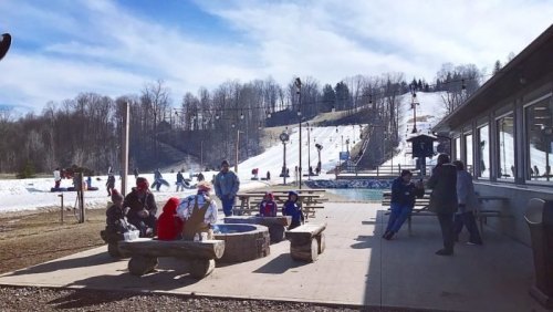 A snowy ski resort scene with people gathered around a fire pit, enjoying the outdoors and slopes in the background.
