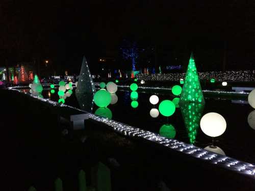 Colorful illuminated decorations in various shapes, including trees and spheres, reflecting in water at night.