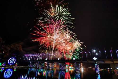 Colorful fireworks burst over a bridge adorned with lights, reflecting in the water below at night.