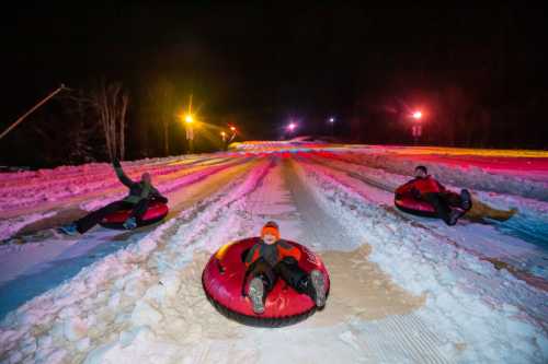 Three people on snow tubes slide down a brightly lit, snowy slope at night.