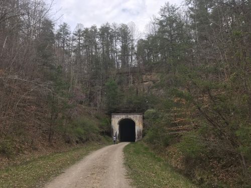 A dirt path leads to a dark tunnel surrounded by trees and rocky cliffs in a wooded area.