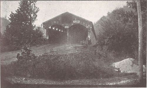 A vintage photo of a covered bridge surrounded by trees and a dirt path, with a foggy atmosphere.