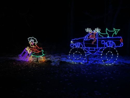 Colorful holiday lights depict a figure in a dress and a decorated truck with reindeer, set against a dark background.