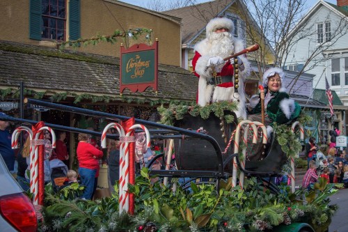 Santa Claus and an elf wave from a decorated sleigh in a festive parade, surrounded by candy canes and holiday cheer.