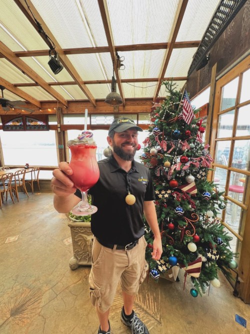 A smiling man in a black shirt holds a colorful drink in front of a decorated Christmas tree indoors.