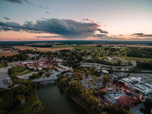Aerial view of a small town by a river, surrounded by fields and trees, with a dramatic sunset sky.