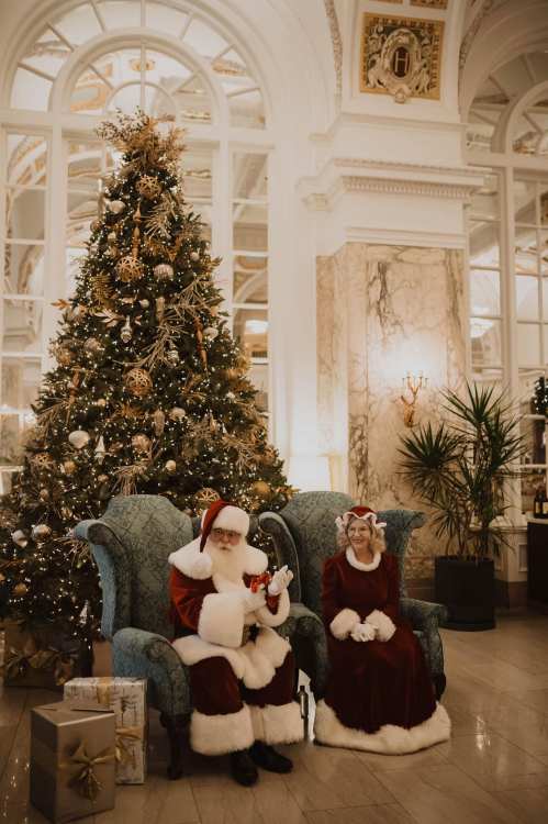 Santa Claus and Mrs. Claus sit together in a festive setting, with a beautifully decorated Christmas tree and gifts nearby.