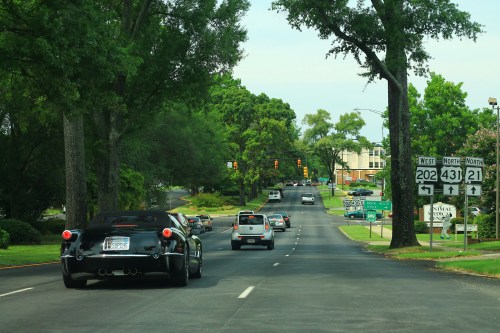 A black sports car drives down a tree-lined road with traffic lights and other vehicles in the background.