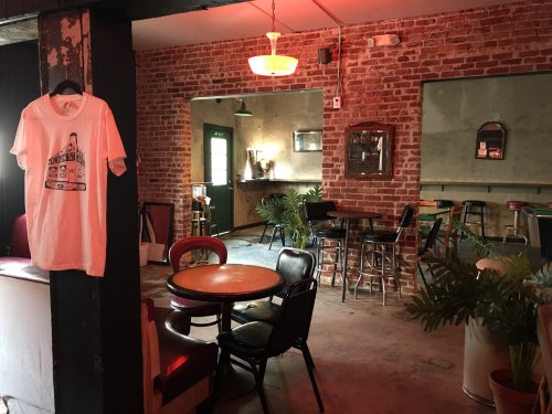 A cozy bar interior with red brick walls, tables, chairs, and a hanging T-shirt on display.