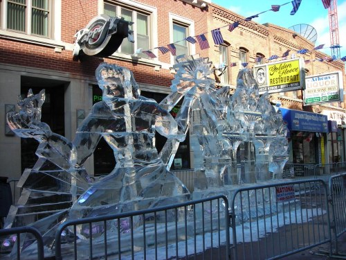A large ice sculpture depicting a figure and intricate designs, set on a city street with shops and flags in the background.