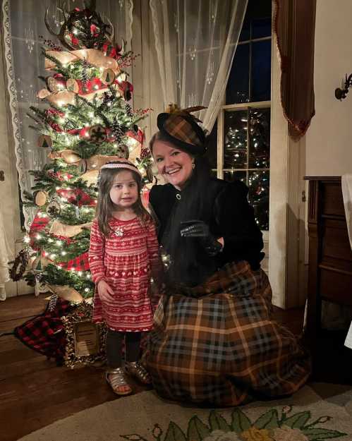 A woman in vintage attire poses with a young girl in front of a decorated Christmas tree, both smiling warmly.