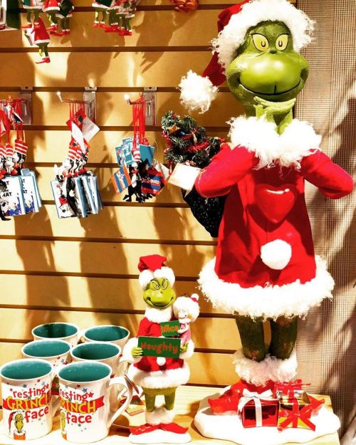 A festive display featuring Grinch figurines in Santa outfits, holiday mugs, and decorations on a wooden shelf.