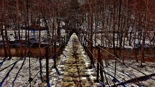 A snowy path leads across a suspension bridge surrounded by bare trees and a flowing stream.