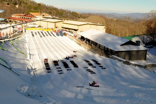 A snowy hill with colorful snow tubes and a lodge in the background, surrounded by trees and mountains.