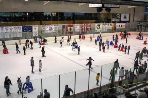 A busy ice skating rink with many people skating and using support aids, surrounded by spectators.