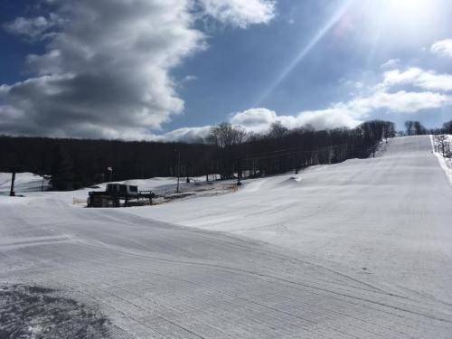 A snowy ski slope under a bright sky with clouds, featuring a grooming machine parked at the base.