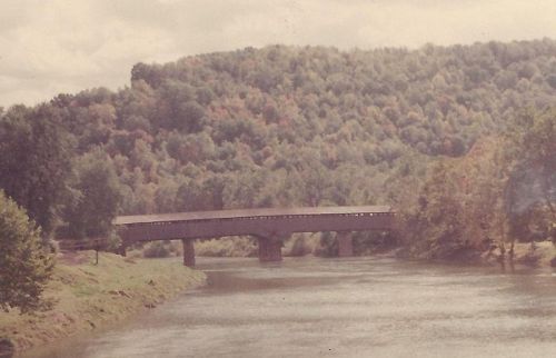 A covered bridge spans a river, surrounded by trees with autumn foliage and rolling hills in the background.