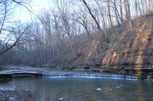 A serene river flows through a wooded area, with rocky banks and trees lining the hillside in soft evening light.