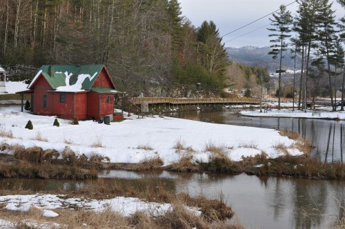 A red cabin with a green roof sits by a snowy riverbank, surrounded by trees and mountains in the background.