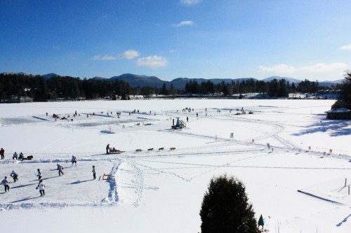 A snowy landscape with people ice skating and dog sledding on a frozen lake, surrounded by mountains and blue skies.