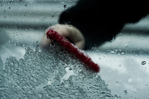 A hand using a red scraper to remove ice and water from a foggy car window.