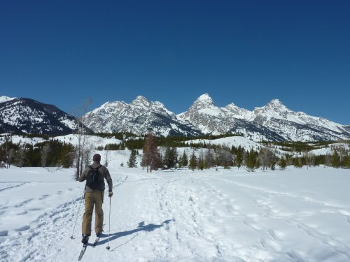 A skier walks on a snowy trail with majestic mountains in the background under a clear blue sky.