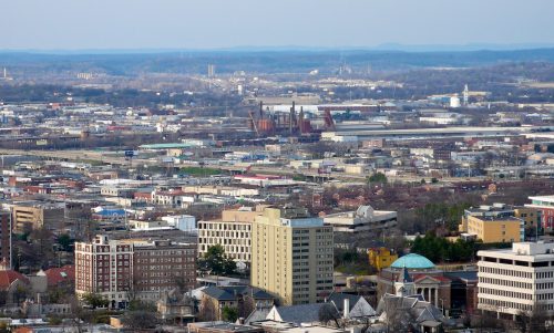A panoramic view of a cityscape featuring buildings, industrial areas, and distant mountains under a clear sky.