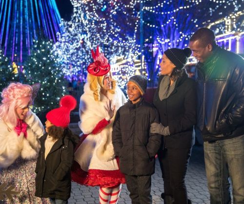 A festive scene with a family and two performers in winter attire, surrounded by colorful holiday lights.