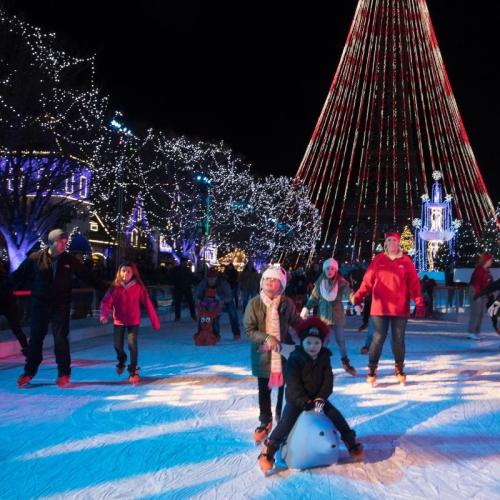 A festive ice skating rink with people skating under colorful lights and a tall Christmas tree in the background.