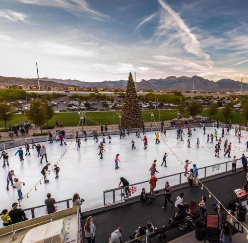 A bustling outdoor ice skating rink with a large Christmas tree, surrounded by mountains and a clear sky.