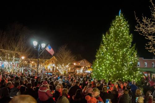 A large crowd gathers around a brightly lit Christmas tree at night, with festive decorations and an American flag in the background.