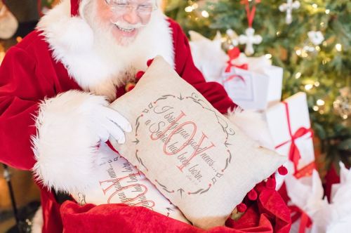 Santa Claus smiles while holding decorative pillows with festive designs, surrounded by Christmas gifts and a tree.
